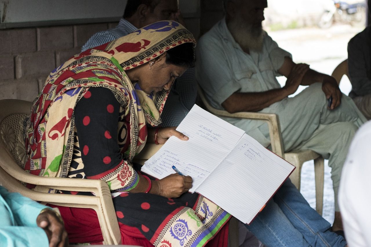A woman sits on a chair, surrounded by people, writing in a notebook on her lap.
