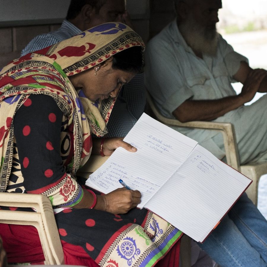 A woman sits on a chair, surrounded by people, writing in a notebook on her lap.