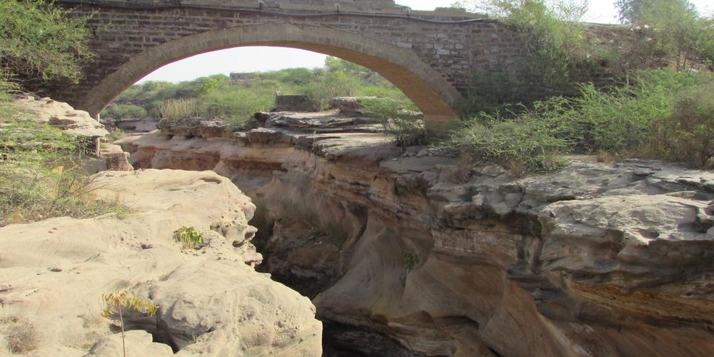An old stone arch bridge spans across a rocky ravine in an arid landscape.