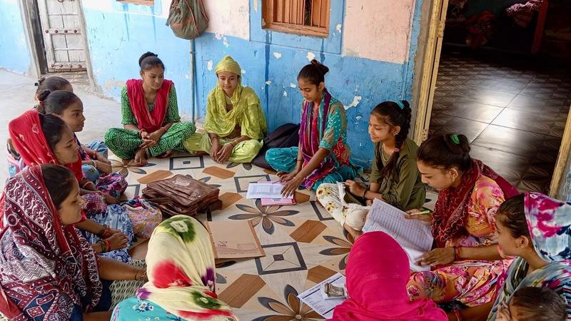Women sit on the floor indoors, near a table and wall. Their diverse clothing and expressions reflect a rich, shared moment.