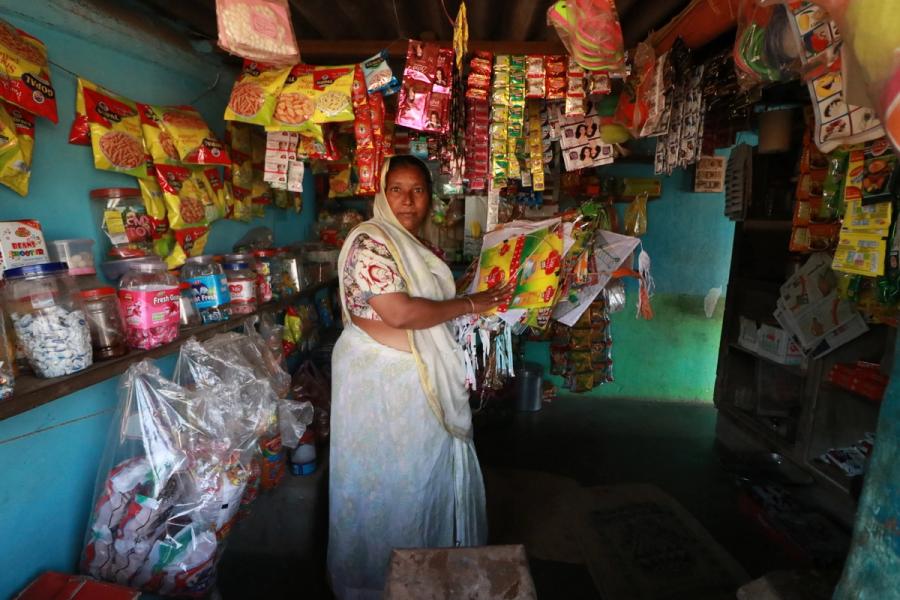 A woman wearing a sari stands inside a small shop filled with various packaged snacks, candies, and goods.