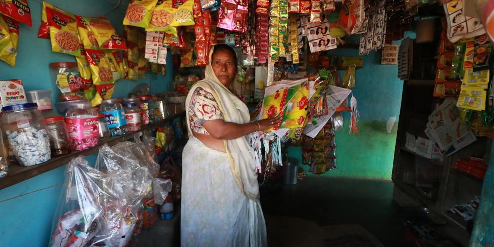 A woman in a white sari stands inside her small village shop with turquoise walls.