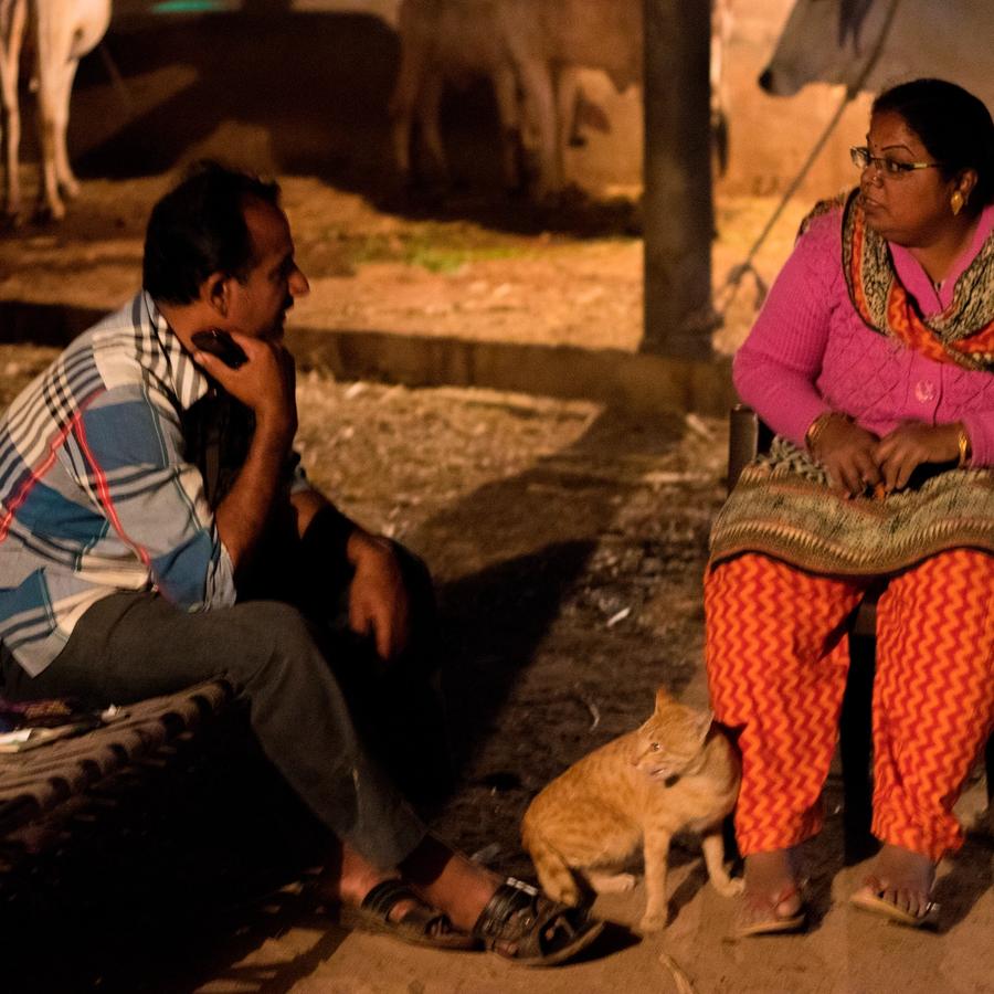 a man sitting on a cot on the left and a woman on a chair on the right side have a conversation