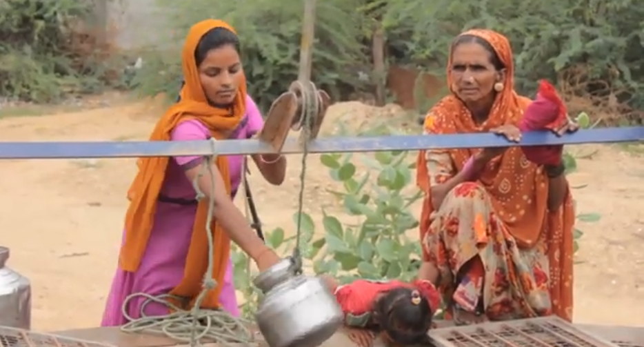 Two women standing near the well, collecting water. one small child looking into well. 