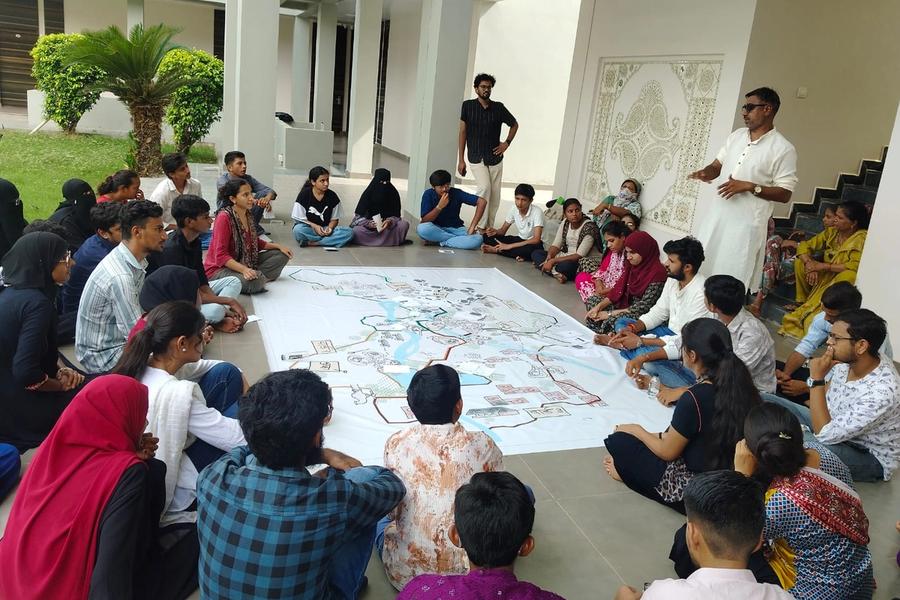 People are sitting in a circle around a big map. A man in a white kurtha standing on the right is addressing the group.