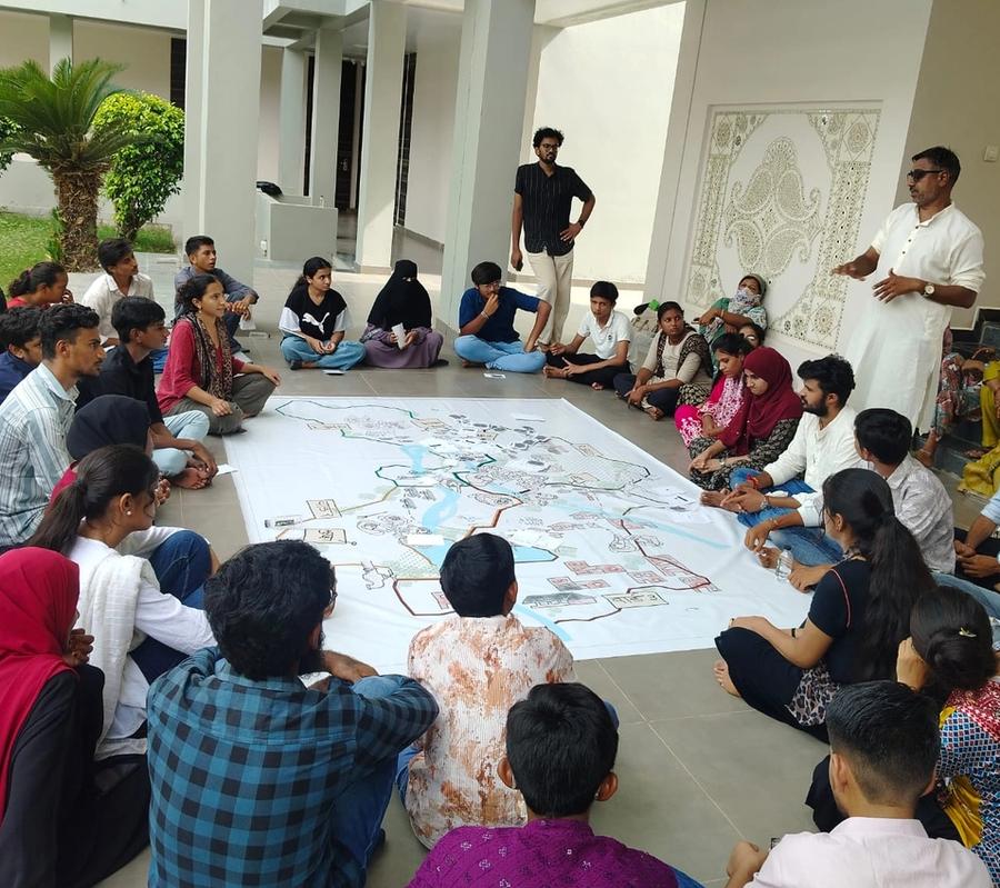 People are sitting in a circle around a big map. A man in a white kurtha standing on the right is addressing the group.