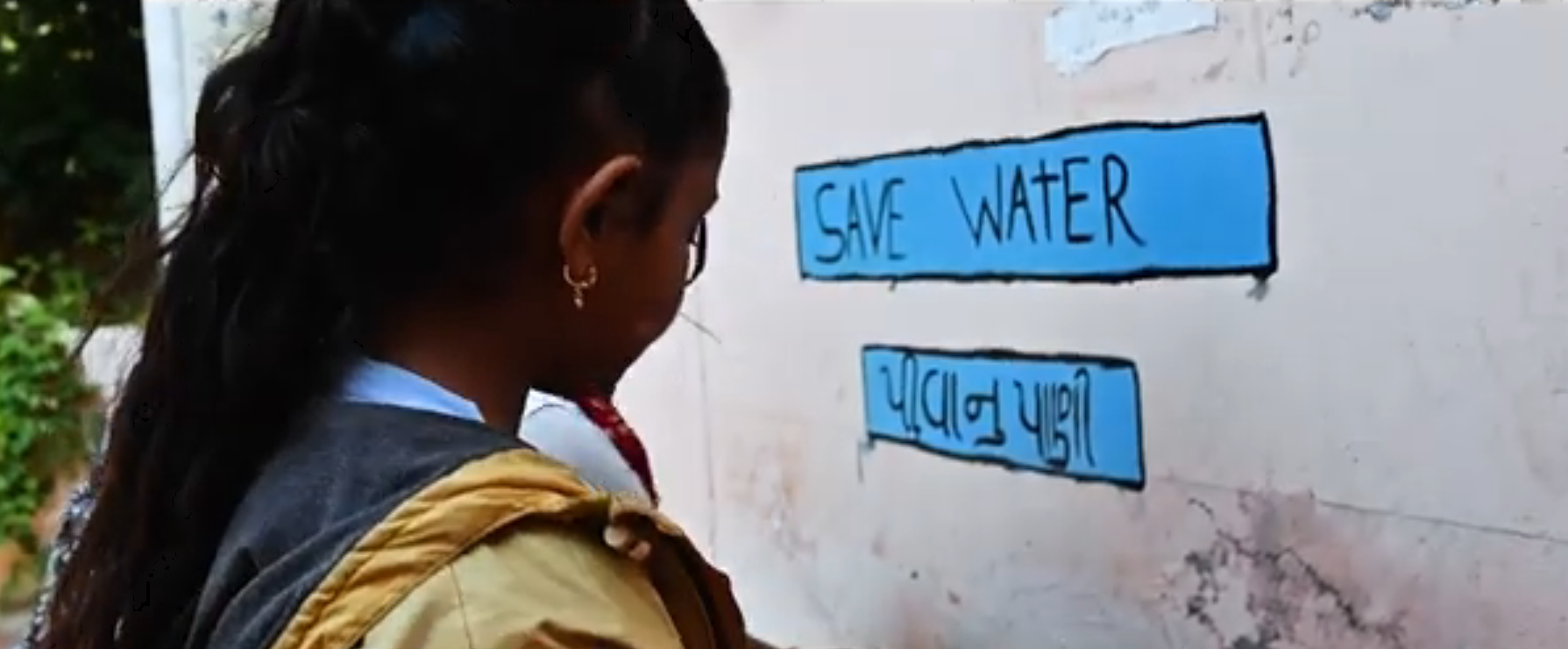 Two young girls washing their hands. A poster written "Save Water" is pasted on the wall. 