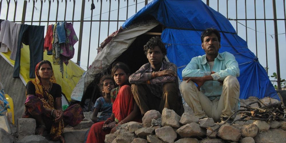 A family sits outside a makeshift shelter constructed with a blue tarpaulin in what appears to be an informal settlement.