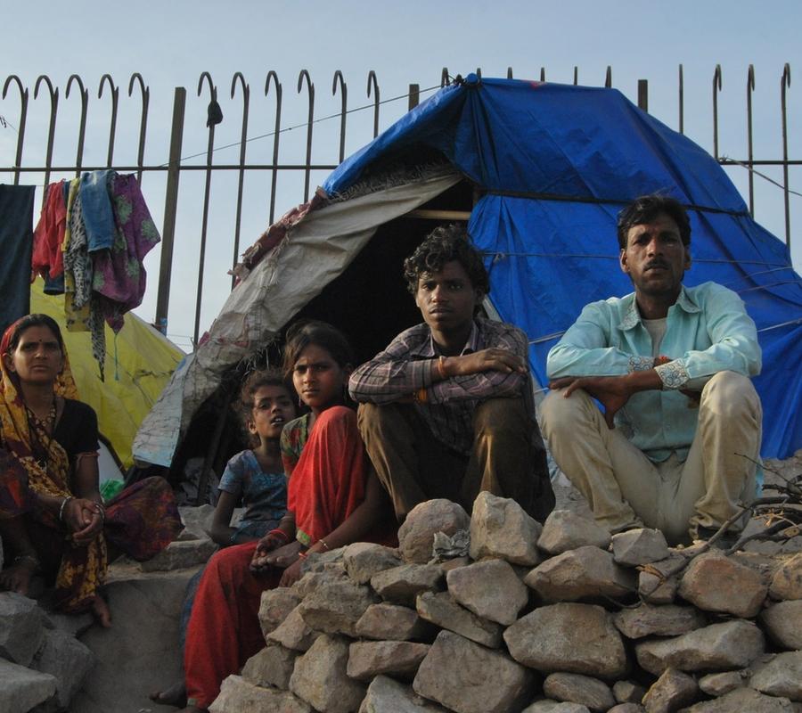 A family sits outside a makeshift shelter constructed with a blue tarpaulin in what appears to be an informal settlement.