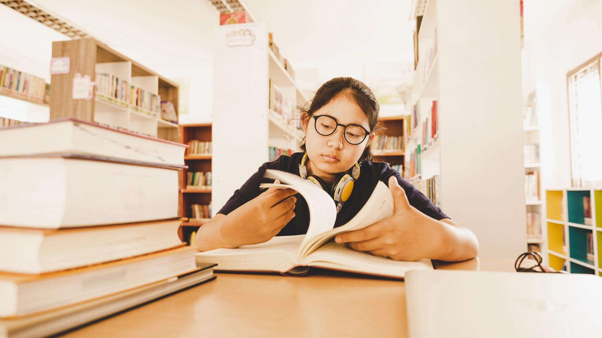 A young student wearing glasses reads a book at a library table, with a stack of books nearby.