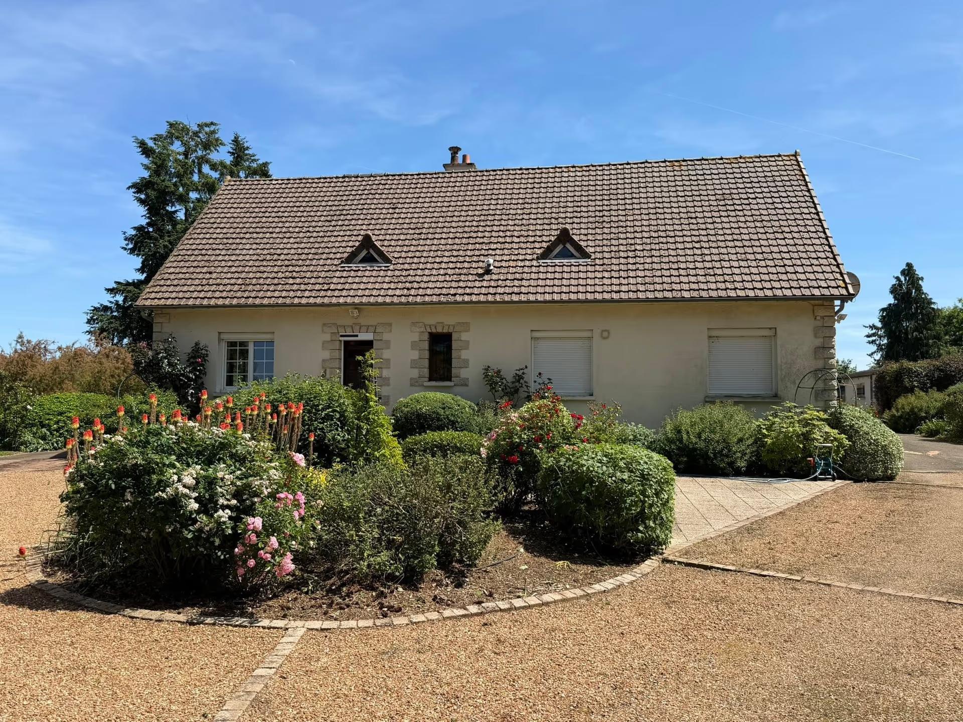 Massifs de rosiers et arbustes taillés en boule devant une longère, au bord d’une cour en gravier