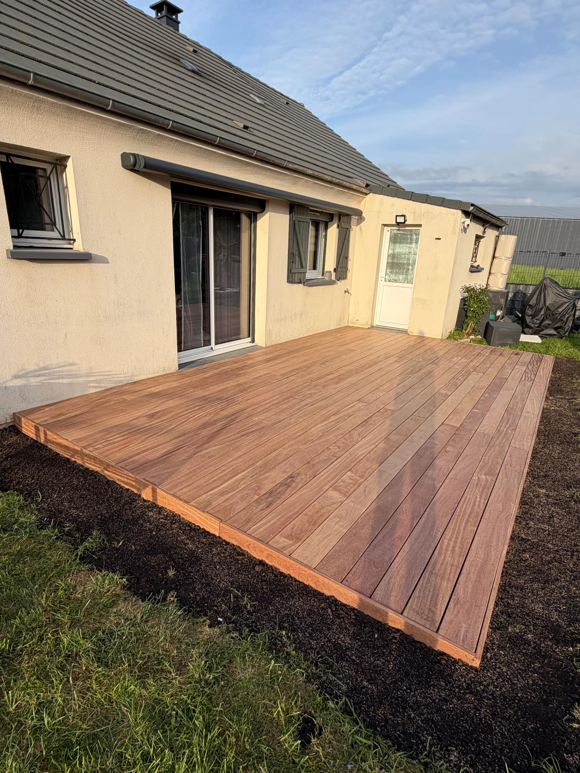 Terrasse en lames de bois exotique Cumaru posée le long d’une maison, bordée de terre fraîchement retournée