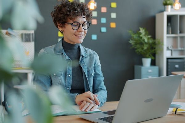 A smiling woman with curly hair works at a laptop.