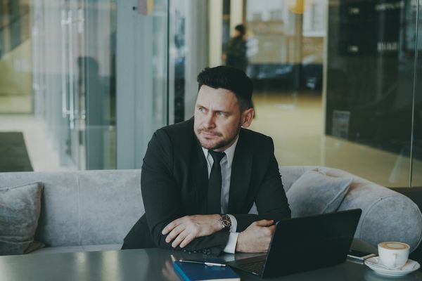 Man in suit sitting at table with laptop and coffee.