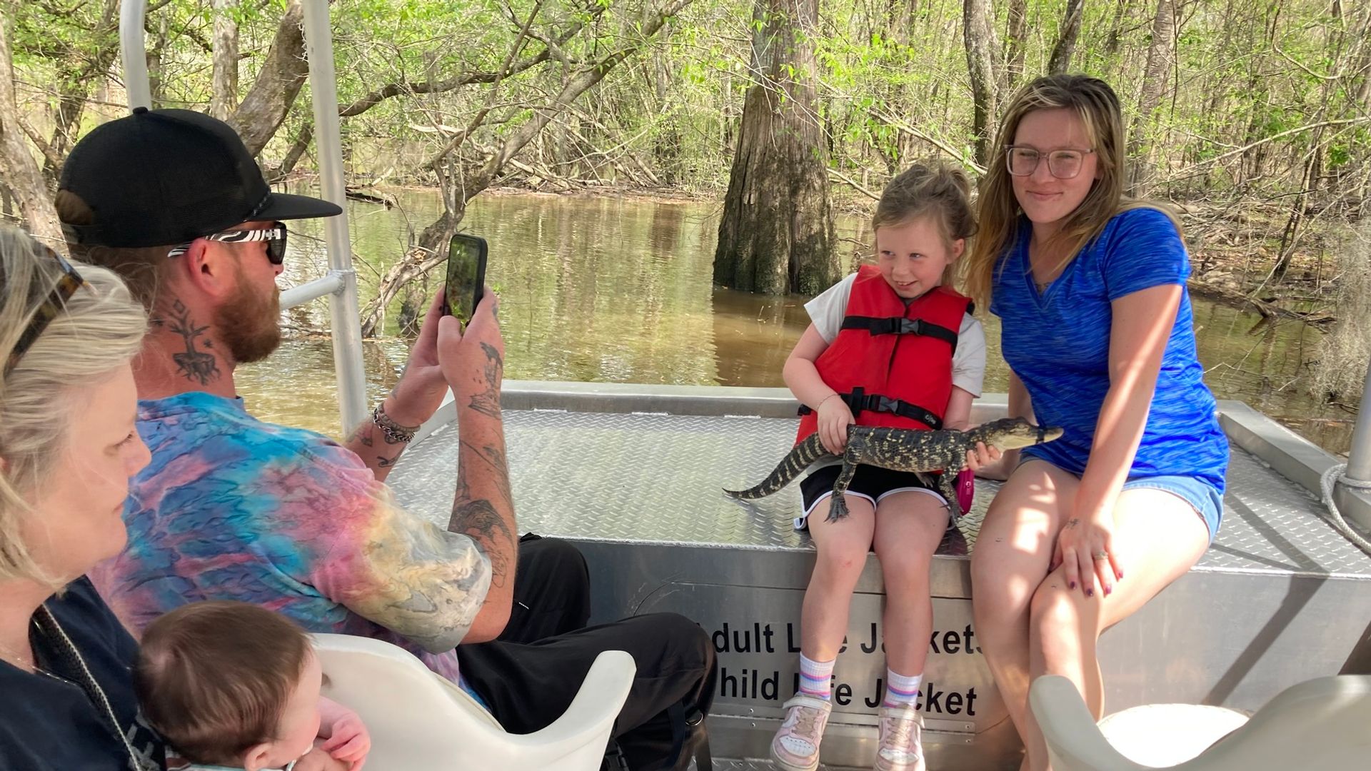 Family enjoying a private Honey Island Swamp tour — daughter holding baby alligator while dad films the moment