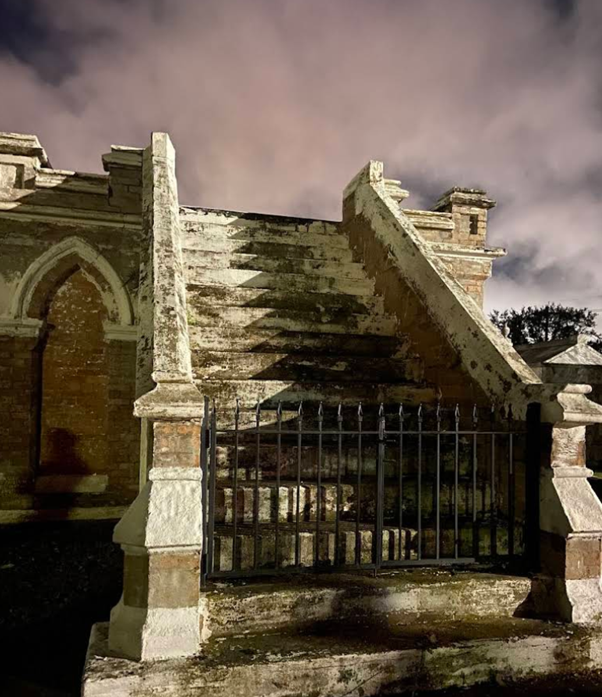 Weathered above-ground staircase tomb behind an iron gate at night