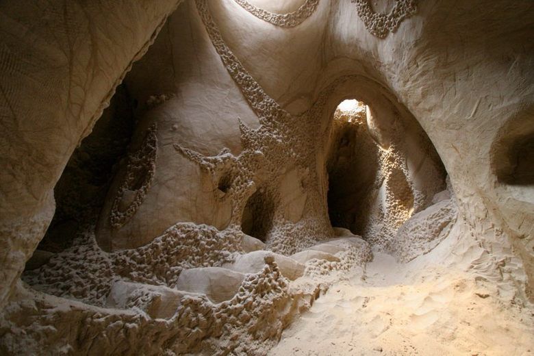 Sunlight illuminates intricate, hand-carved patterns and smooth arches inside a subterranean sandstone cave chamber.