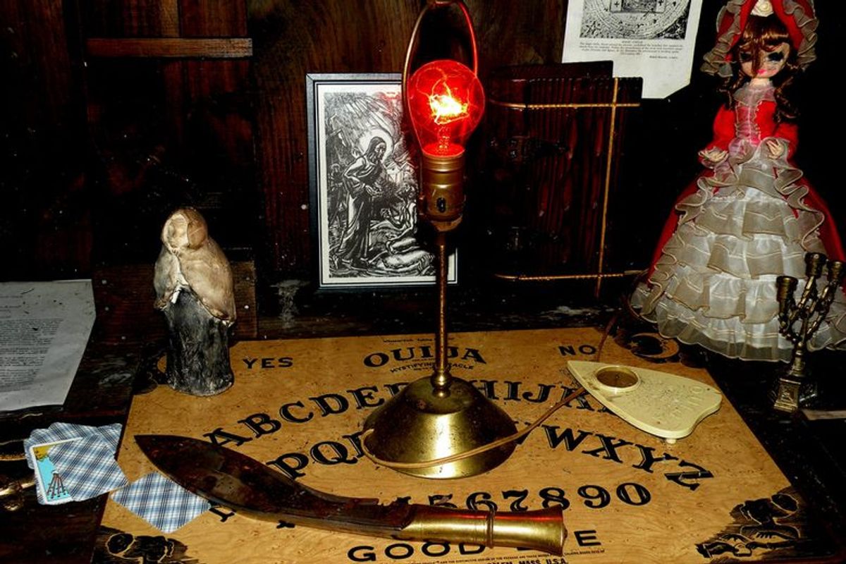 A red light bulb illuminates a table with a Ouija board, a large curved knife, tarot cards, and a Victorian doll.