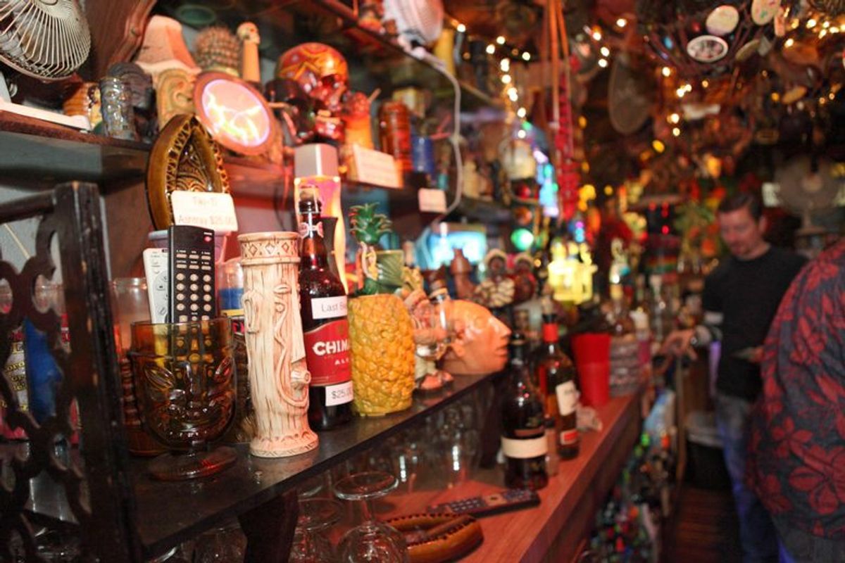 A crowded bar shelf at Tiki-Ti filled with tiki mugs, pineapple decor, liquor bottles, and eclectic memorabilia.