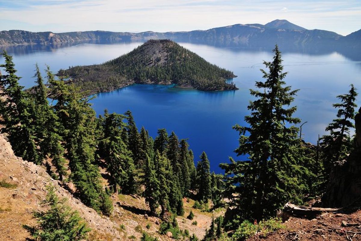 A deep blue lake fills a volcanic caldera with a conical, tree-covered island at its center and evergreen trees in the foreground.