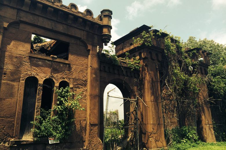 The crumbling brownstone gatehouse of Mount Moriah Cemetery is overgrown with vines and greenery under a bright sky.