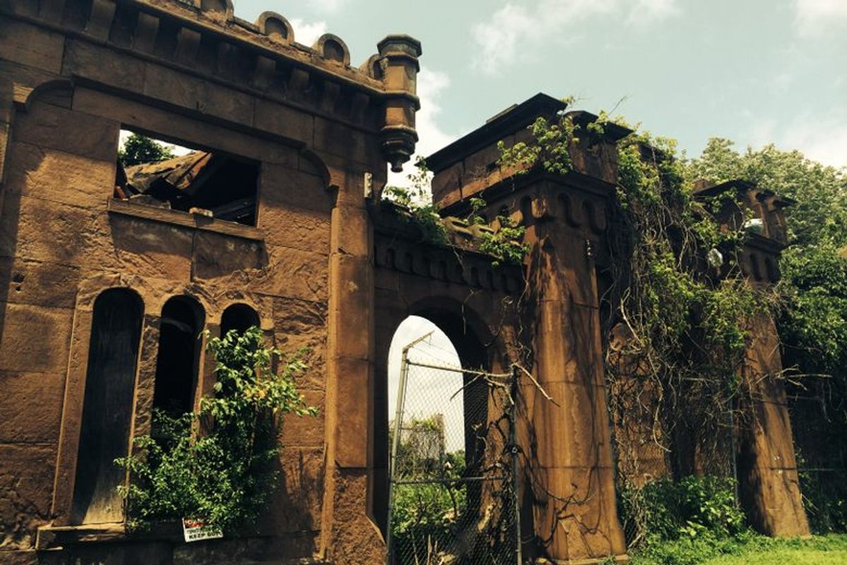 The crumbling brownstone gatehouse of Mount Moriah Cemetery is overgrown with vines and greenery under a bright sky.