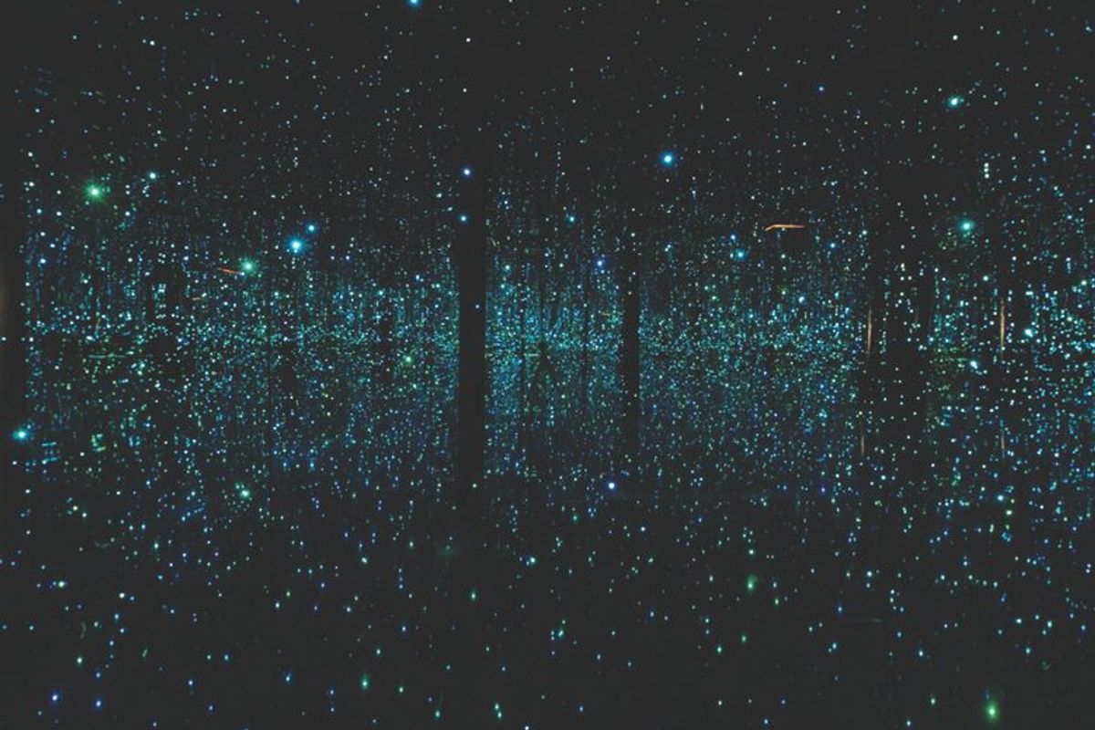 Tiny blue and green lights reflect infinitely in a dark mirror room, creating the appearance of a starry night sky.
