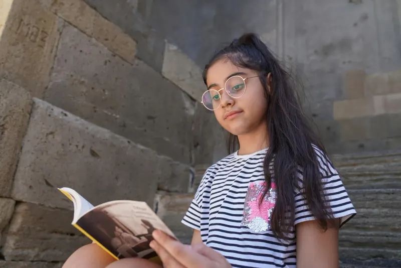 A young girl with glasses enjoys reading a book while sitting on stone steps, surrounded by rustic walls.