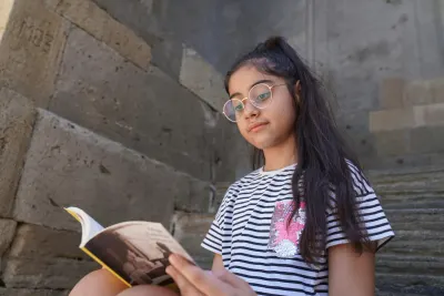 A young girl with glasses enjoys reading a book while sitting on stone steps, surrounded by rustic walls.