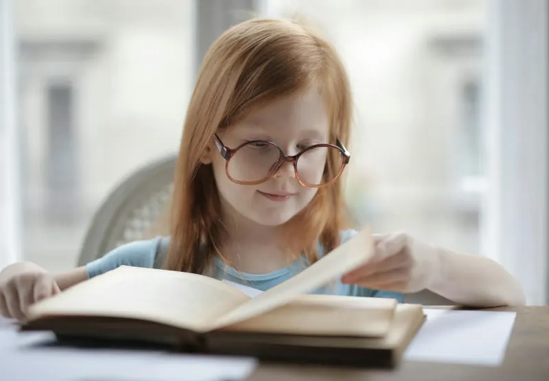 A young girl with eyeglasses reading a book at a desk indoors, focused and concentrated.