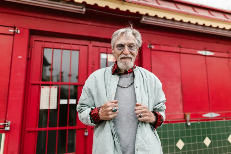 Elderly man with stylish outfit smiling confidently in front of a red building exterior.
