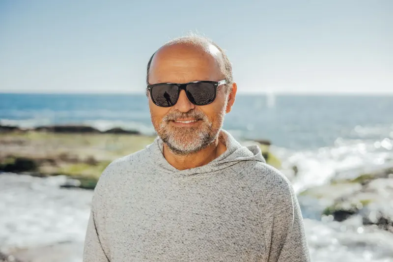 Senior man wearing sunglasses, smiling at the beach on a sunny day.