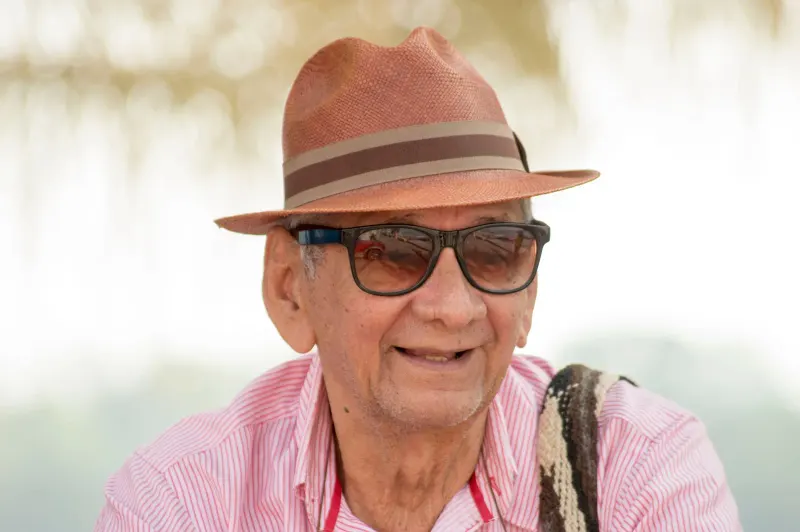 Elderly man wearing a hat and sunglasses smiling warmly outdoors.