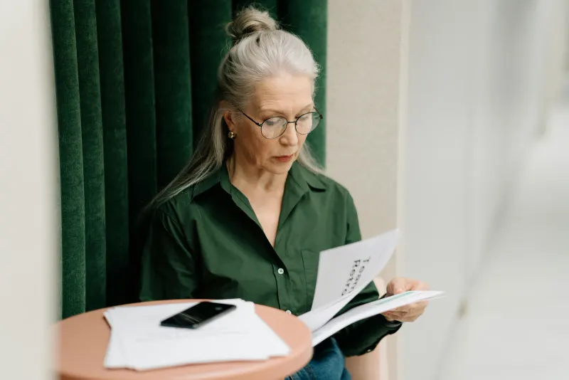 Elderly woman with gray hair reading papers at a small table indoors, focused and thoughtful.