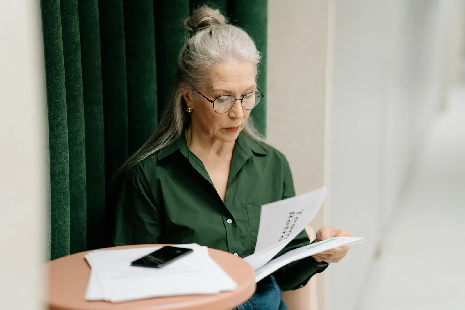 Elderly woman with gray hair reading papers at a small table indoors, focused and thoughtful.