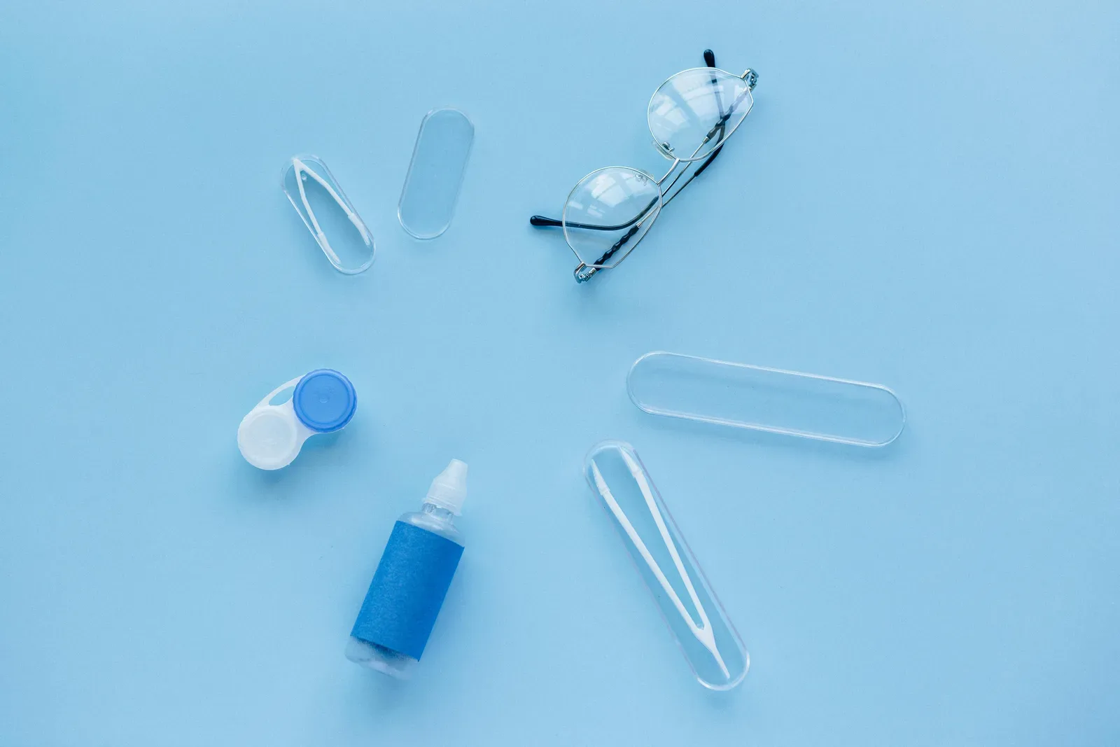 Flat lay of contact lens case, solution, and glasses on a blue background.