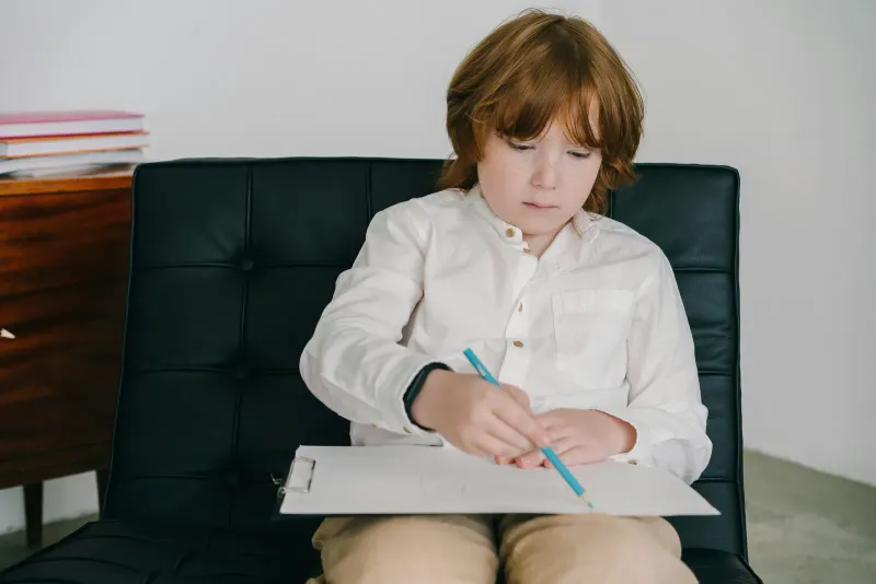 Child with red hair concentrates on drawing with colored pencils while sitting on a black chair.