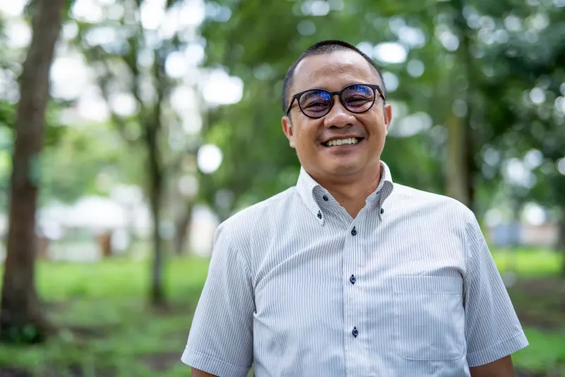 Portrait of a smiling Asian man with eyeglasses standing in a green park setting.