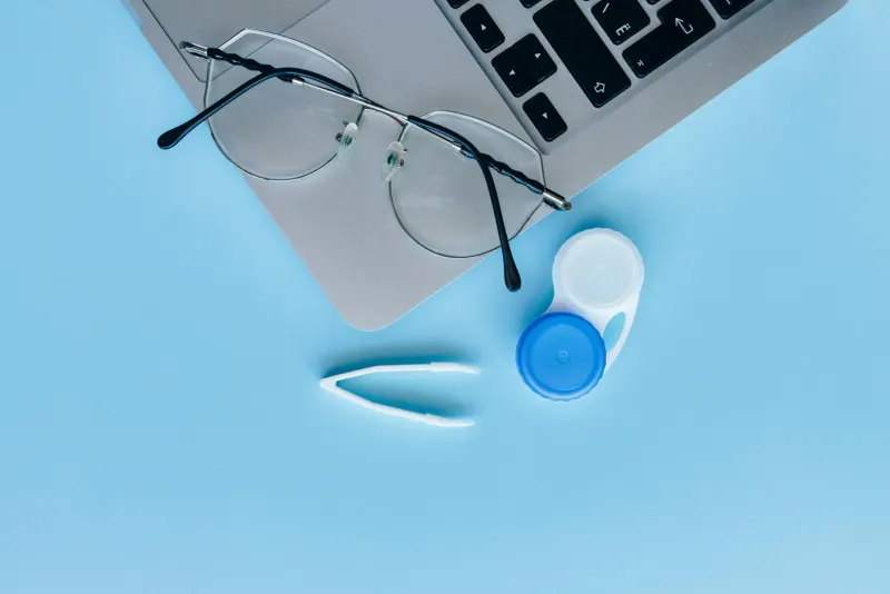 Flat lay of eyeglasses, contact lens case, and laptop on a blue surface.