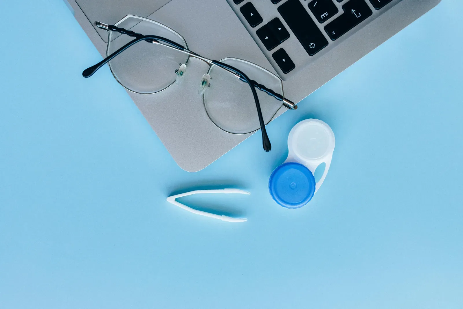 Flat lay of eyeglasses, contact lens case, and laptop on a blue surface.