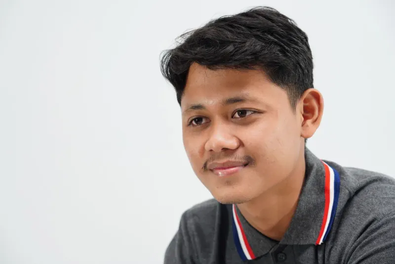 A close-up portrait of a young man smiling, wearing a gray polo shirt with a white background.