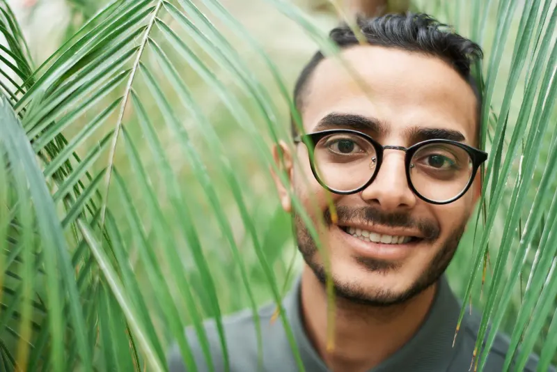 Close-up portrait of a happy man with eyeglasses among lush green palm leaves, expressing joy and nature connection.