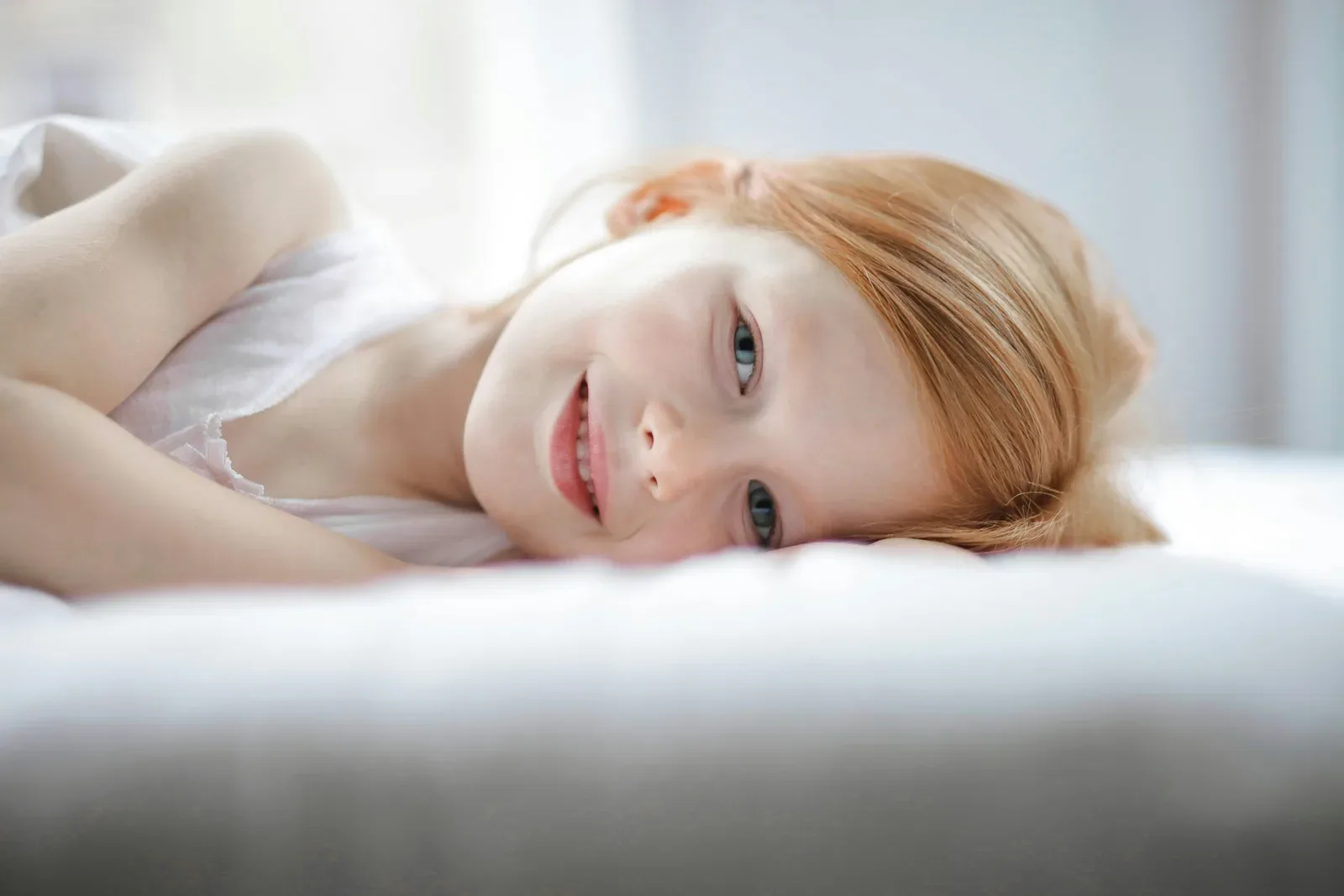 A young girl with red hair smiling while lying on a bed indoors, conveying comfort and joy.
