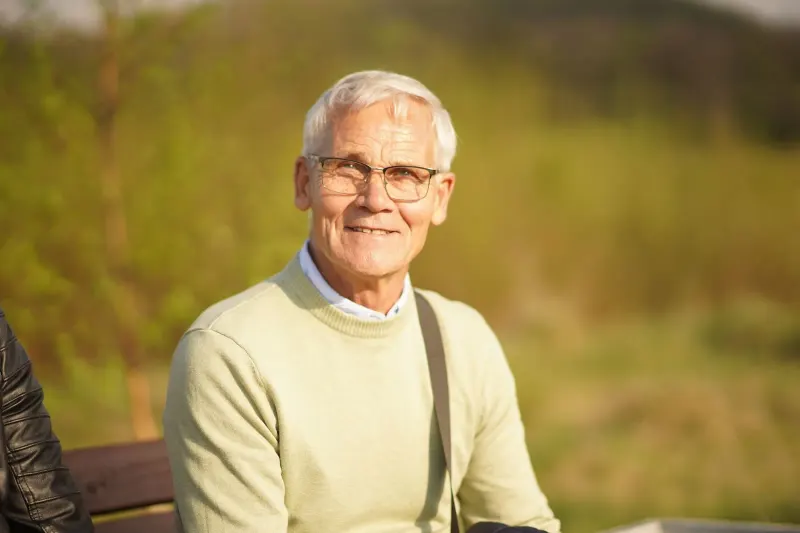 Senior man smiling warmly while sitting outdoors in bright sunlight, wearing glasses.