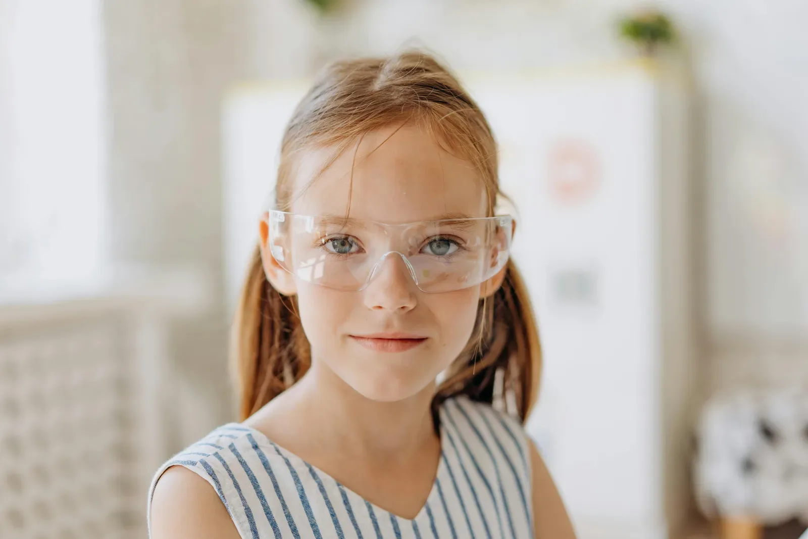 A young girl in a striped dress and safety glasses standing in a bright classroom.