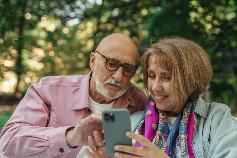 Happy senior couple using smartphone in a park, enjoying sunny outdoors.