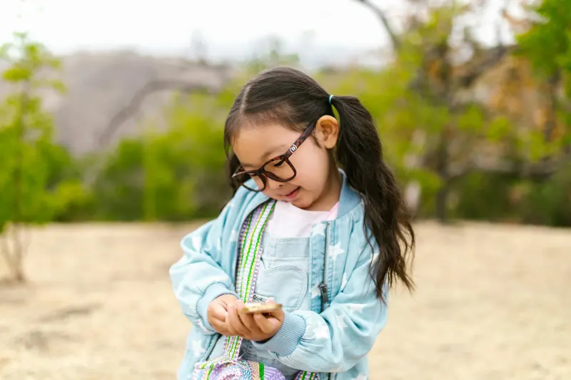 Curious young girl in glasses looking at a compass in a sunny outdoor setting.