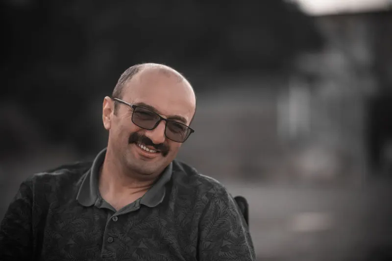 Man with eyeglasses smiles warmly outdoors, wearing a gray polo shirt.