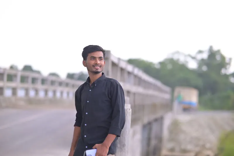 Young man in black shirt smiling and standing on a bridge on a bright day.