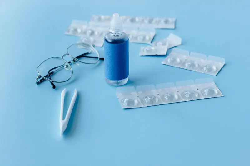 Close-up of contact lens solution bottle with eyeglasses and tweezers on blue backdrop.